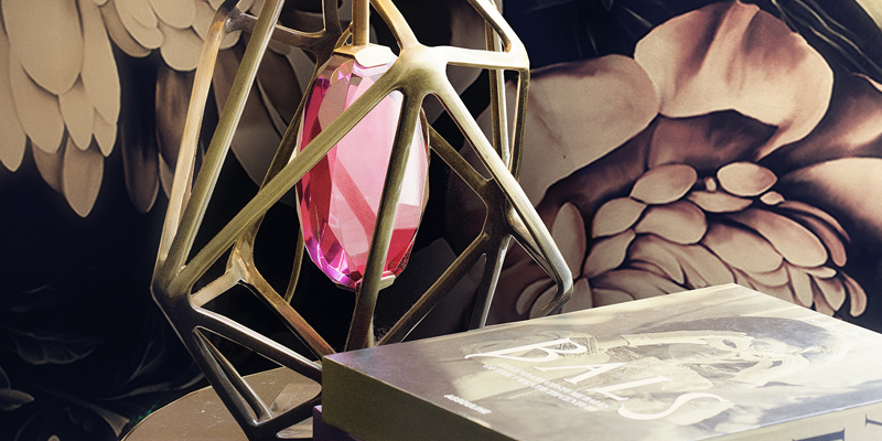 A pink Gem Table Lamp inside a gold geometric frame, resting on a table with a book, against a floral backdrop.