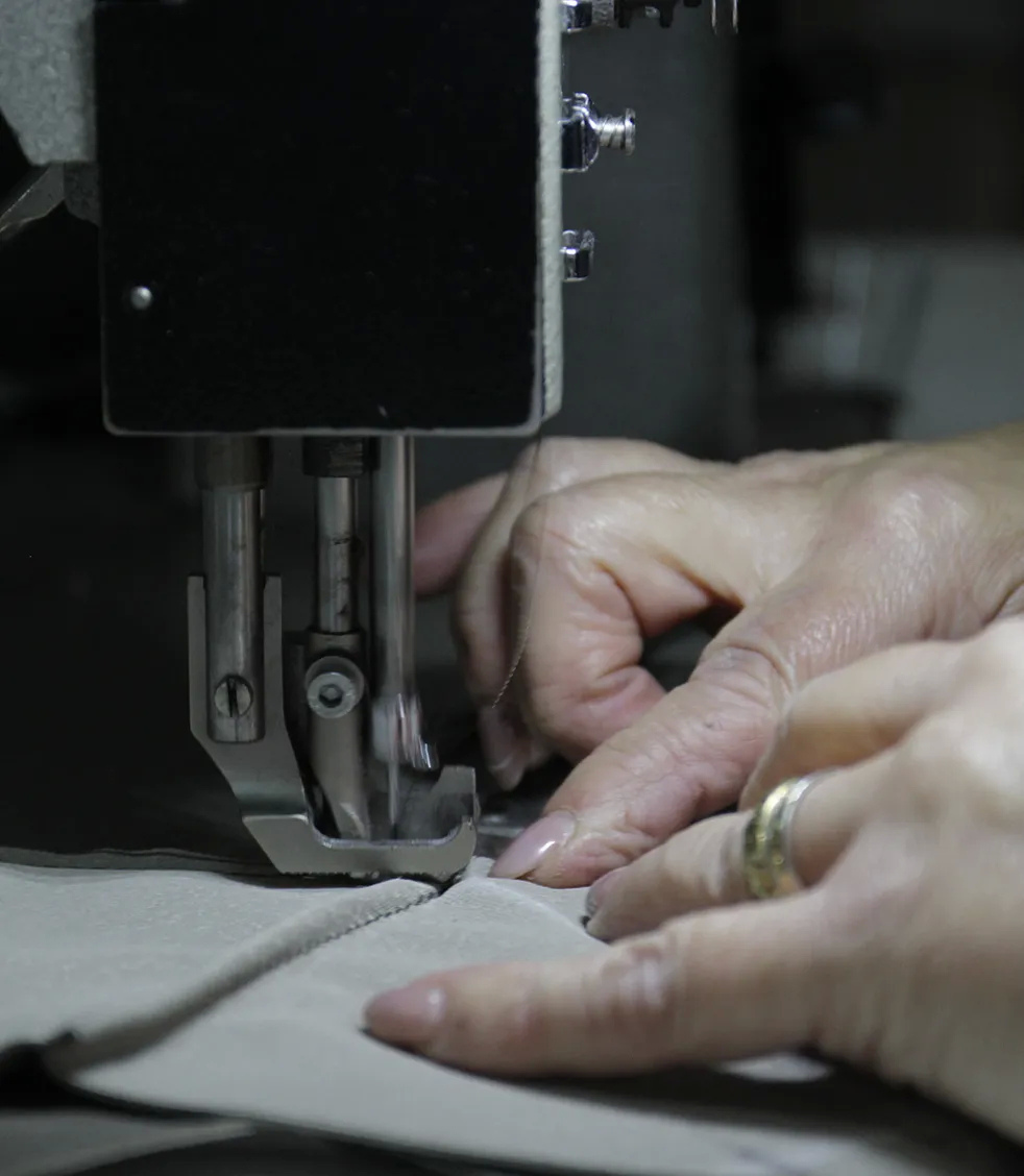 Close-up of a person's hands guiding light-colored fabric under the needle of a sewing machine, illustrating the meticulous craftsmanship behind luxury furniture.