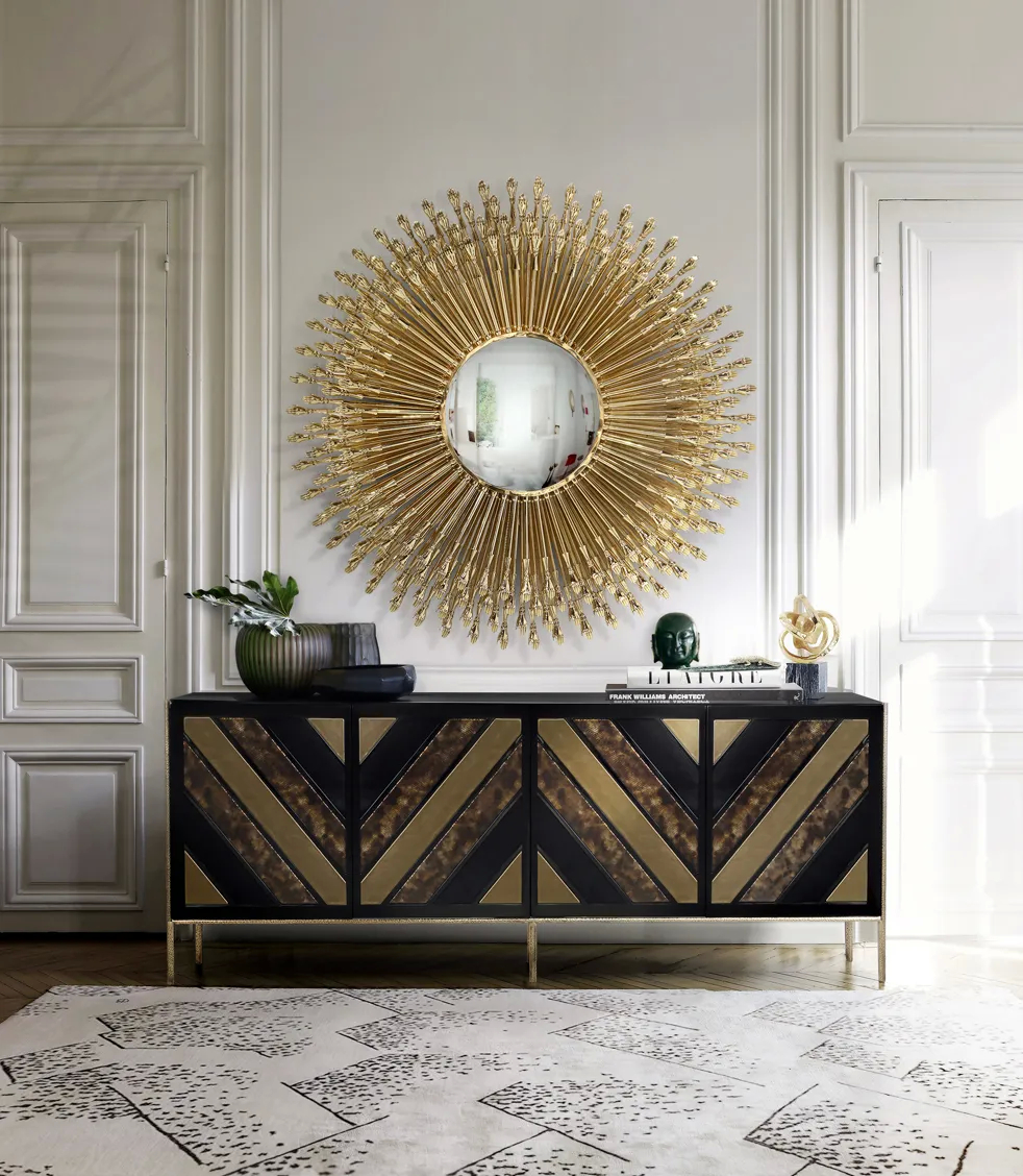 The black and gold Opium Cabinet by KOKET is displayed beneath a radiant gold mirror, adding a touch of luxury to the room.