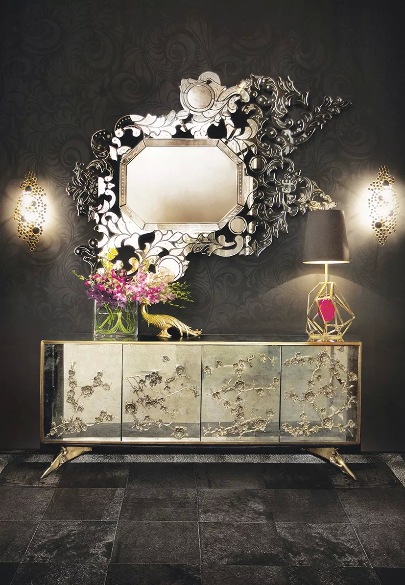 This hallway exudes an air of luxurious elegance, as though inviting you into a realm of timeless beauty. Anchoring the space is a silver Spellbound Cabinet, serves as a stunning focal point, adorned with intricate floral motifs, evoking the fragile beauty of nature. Above it, the breathtaking Addicta Mirror unfolds in an elaborate frame, its edges catching the light. The Gem Table Lamp, with a geometric frame and magenta gemstone adds a touch of sophistication, while the Eternity Sconces cast a soft flow, creating an atmosphere of quiet luxury.