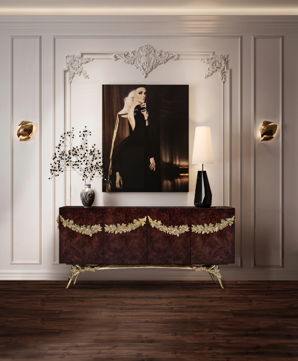 Luxurious sideboard in walnut root veneer with ornate gold-leaf garlands and sculpted gold legs, styled against a white paneled wall with a modern portrait, sculptural lamp, and decorative vase.