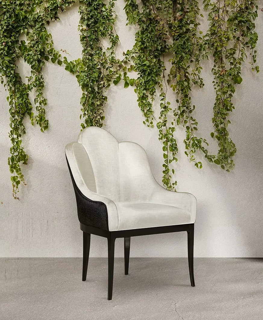 A sophisticated Anastasia Dining Chair by Koket with a white velvet back and dark wood legs sits against a green ivy wall.
