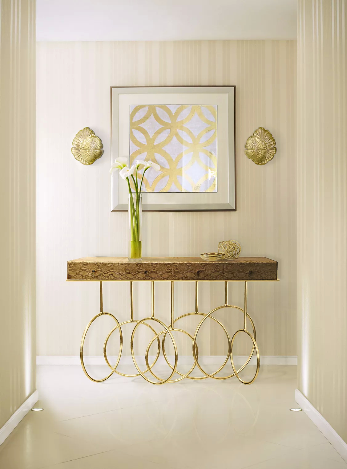 In bright hues, this hallway features a Burlesque Console, with its unique design, a textured top and a golden base with interlocking circles. Above it, there is an artistic feature surrounded by two Passion Sconce with a simple yet elegant design resembling leaves.