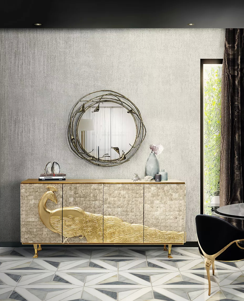 This dining room is a masterpiece of understated luxury. The walls have a textured soft tone. At the room’s heart, an outstanding Camilia Cabinet graces the space, adorned with a golden peacock, its intricate feathers unfolding across the cabinet. Above, a round Stella Mirror framed in entwined copper calla lilies and stems reflects the room's light. The Chandra Dining Chair, demonstrating its distinctive feature of only having 3 legs, contrasting with the space’s soft hues, with its black velvety upholstery.
