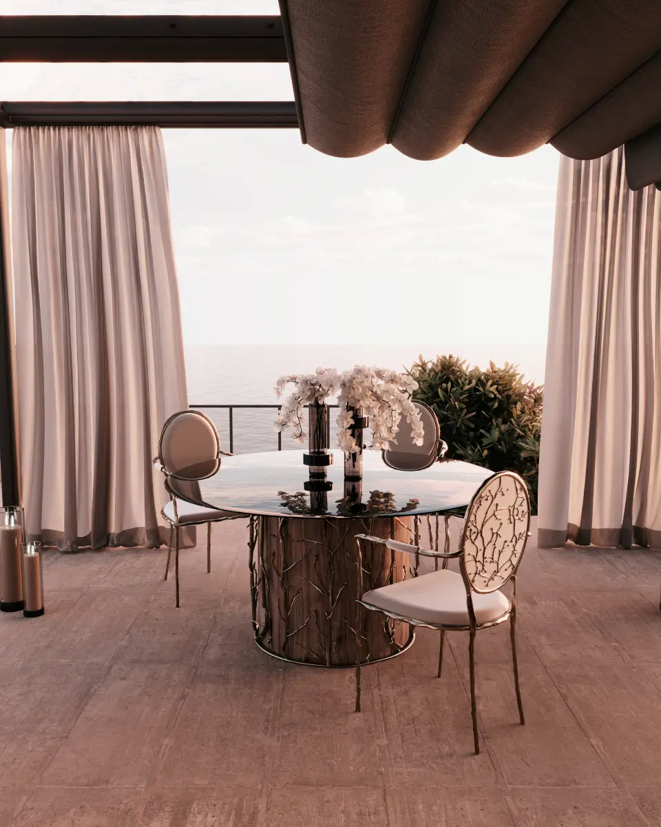Elegant outdoor dining area featuring Koket’s Enchanted Dining Table with a glass top and intricate branch-like base, surrounded by matching Enchanted Dining Chairs with metallic frames and white cushions, set under a pergola with flowing curtains and a view of the ocean.