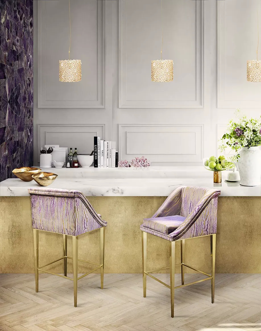 Two purple and gold Geisha Bar Stools by Koket stand at a white marble kitchen island with gold accents and pendant lights.