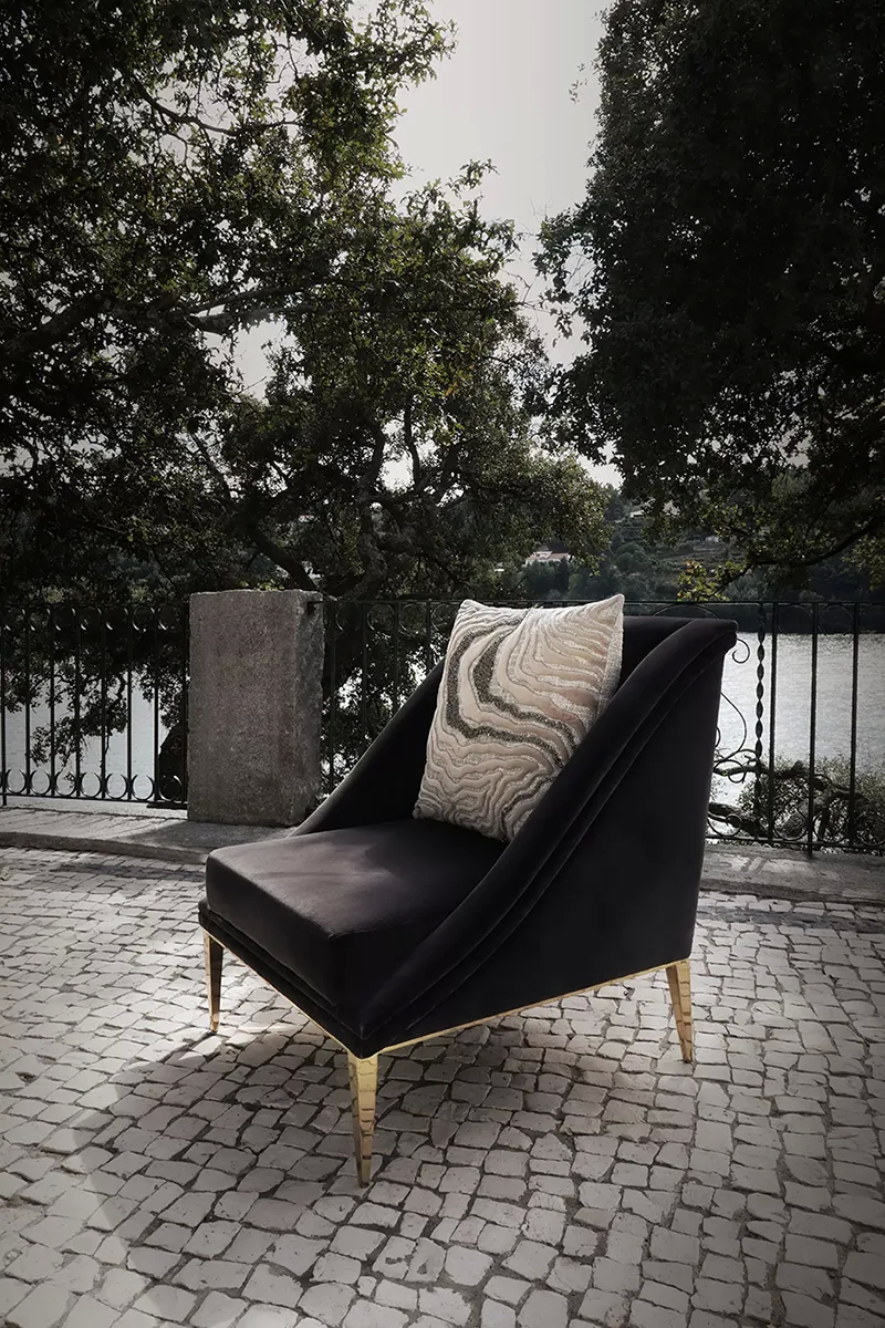 A luxurious black velvet Geisha Chair by Koket with a patterned pillow sits on a stone patio overlooking a lake and trees.