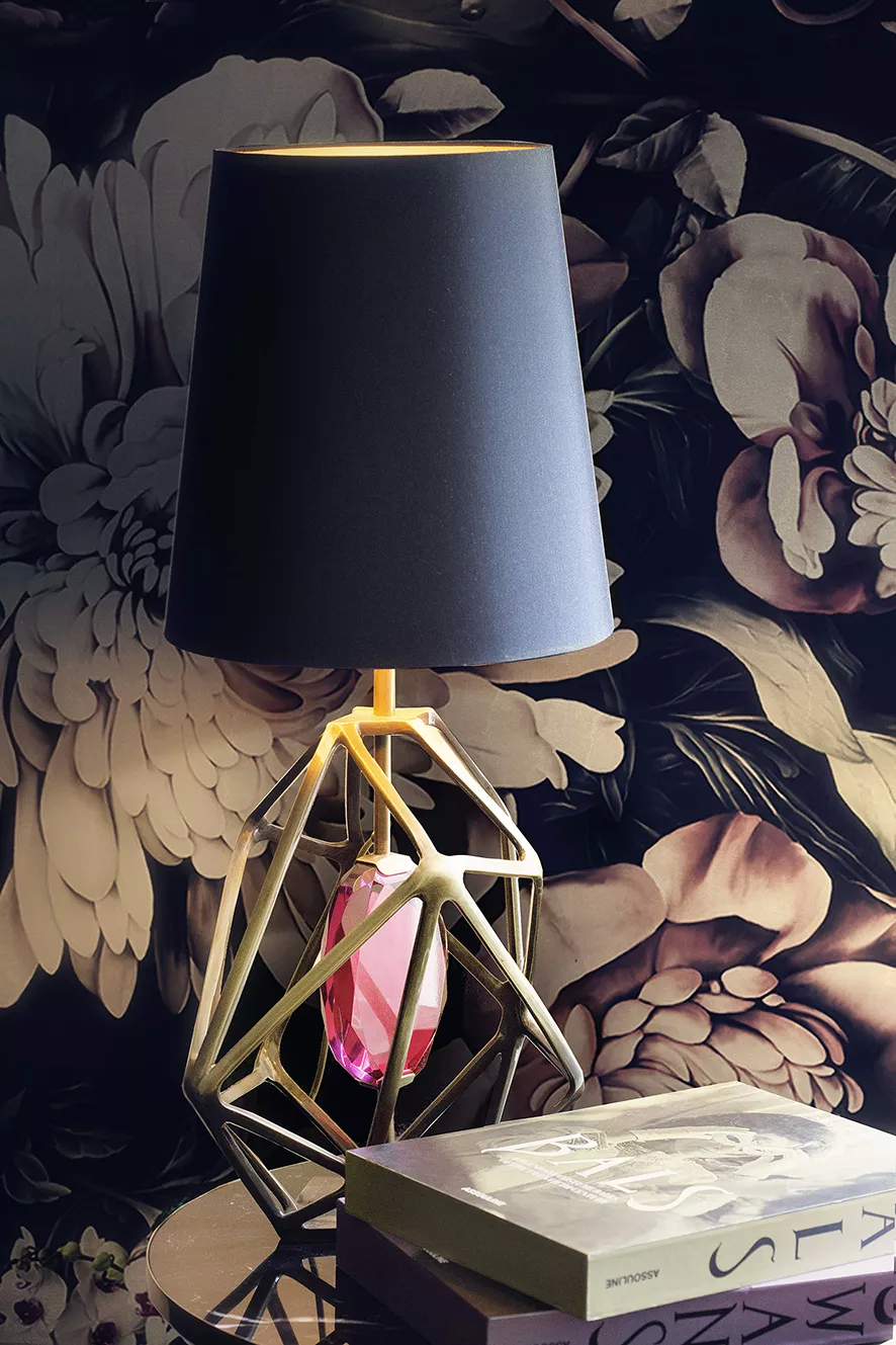 A table lamp with a black lamp shade and an aged brass structure with a blue gem inside, adding a touch of luxury. Next to the table lamp there are books. The background has a floral motif.