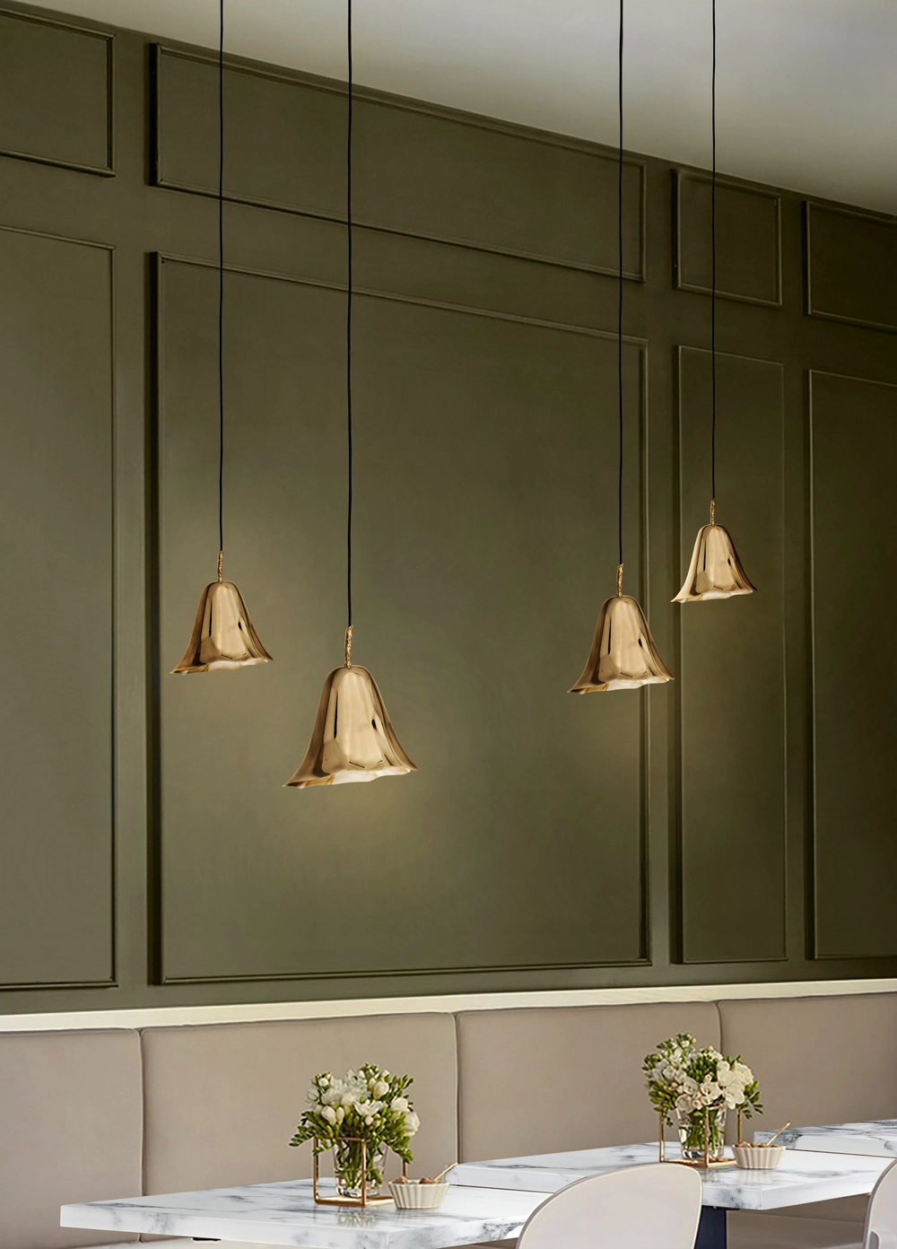 Four bell-shaped gold pendant lights hang from the ceiling against a dark green paneled wall above white marble-top tables with floral centerpieces and beige banquette seating in a modern dining room.