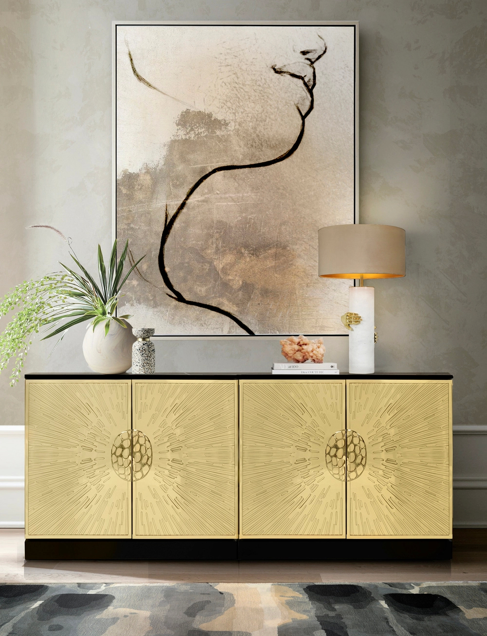 A luxurious Heive Cabinet, a gold console table with sunburst-patterned doors, stands on a dark floor in front of a light gray wall. On top of the cabinet, there's a white vase with greenery, decorative objects, and a Vengeance Table Lamp. Above the cabinet hangs a large abstract painting with a prominent black line forming a profile on a neutral background.