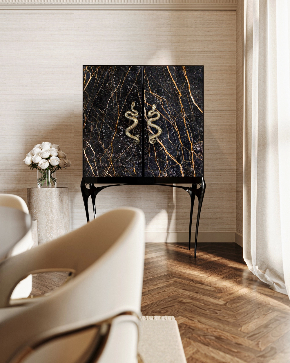 A luxurious black marble cabinet with distinct gold veined patterns and ornate serpent-shaped handles. The modern cabinet stands on elegant black legs against a textured light wallpaper, with a cream chair and white flowers in the foreground.
