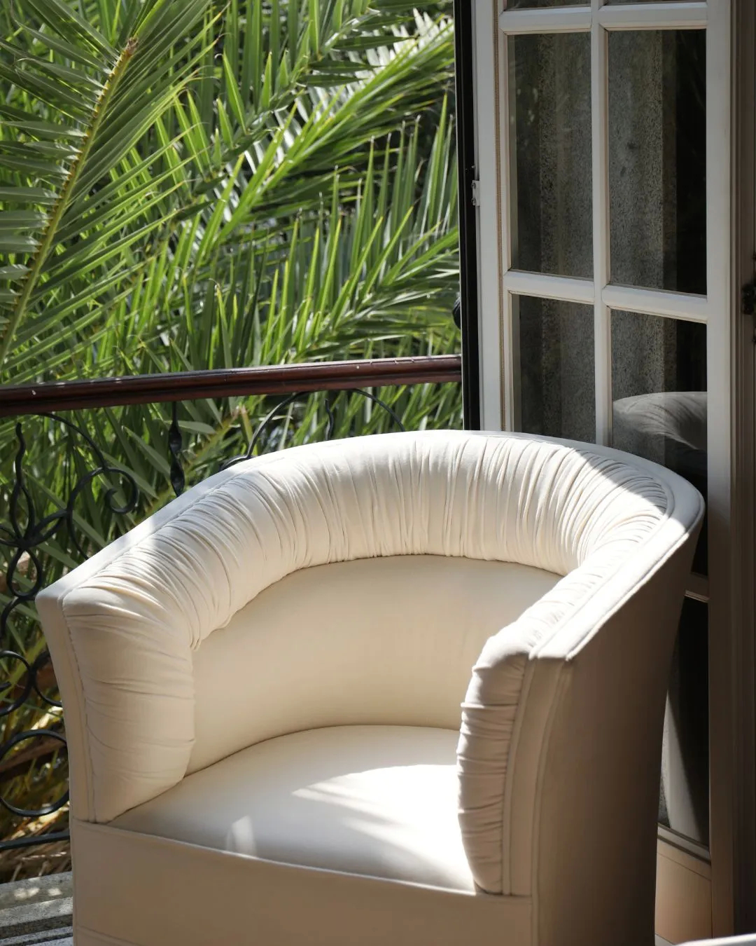 Cream-colored armchair with ruched backrest and curved silhouette placed near a balcony with lush greenery.