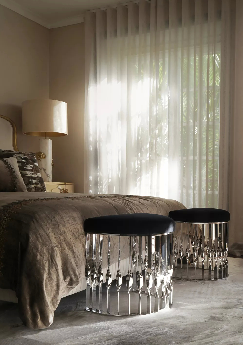 In a neutral-colored bedroom, bathed in natural light, the two Mandy Stools with luxurious soft black velvet upholstery and a silver twisted base, stand at the bottom of the bed. Next to the bed, the Vengeance Table Lamp in cream hues, graces the room with its elegance.