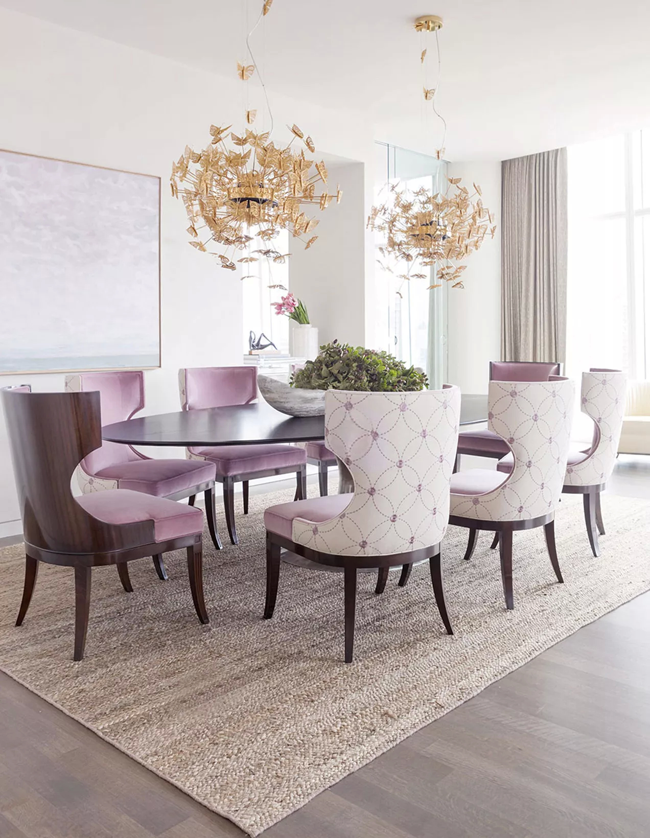 A spacious dining room with a gracious ambiance, where a large table is surrounded by various pink dining chairs. Above it, there are two golden Eternity Chandeliers, inspired by the delicacy of butterflies.