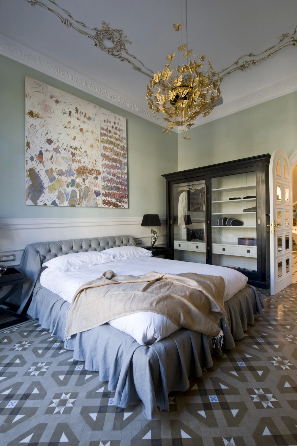 A spacious bedroom with a large bed, a dark wood wardrobe, and a golden Nymph Chandelier, featuring an ornate ceiling and patterned floor.