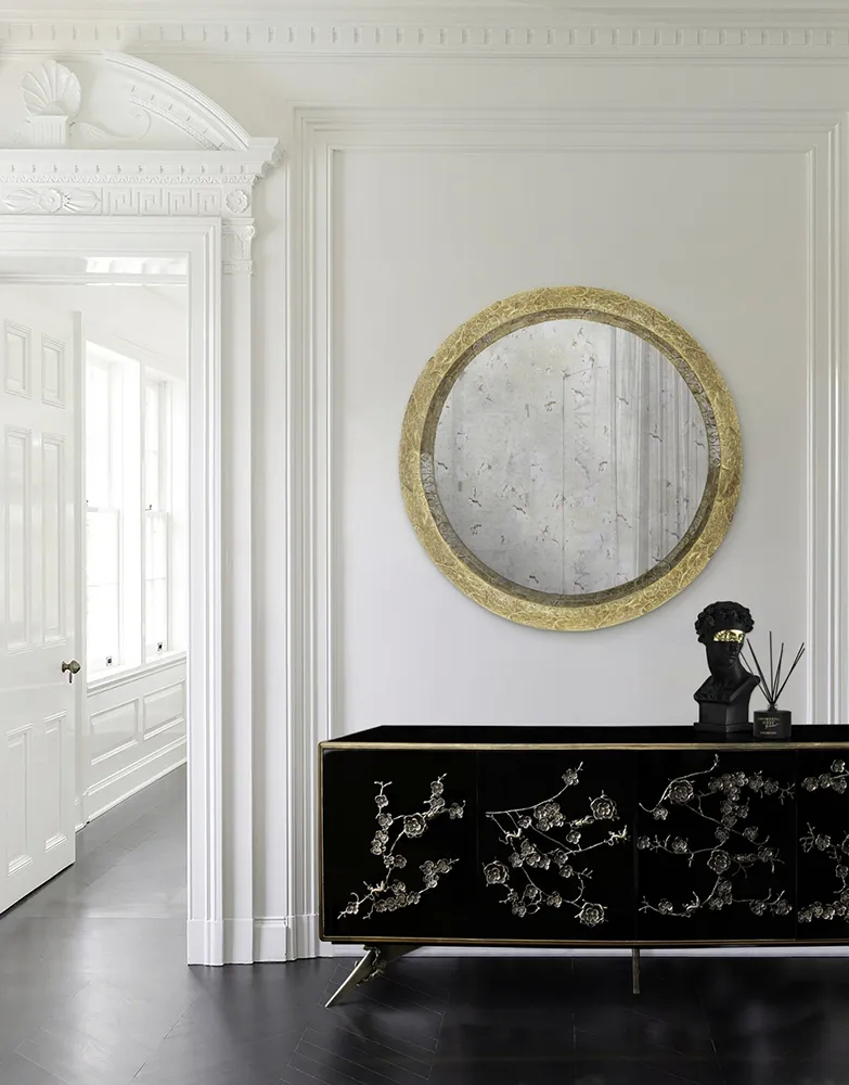 A luxurious black Spellbound Cabinet with gold floral details under a round gold mirror, next to an open white doorway.