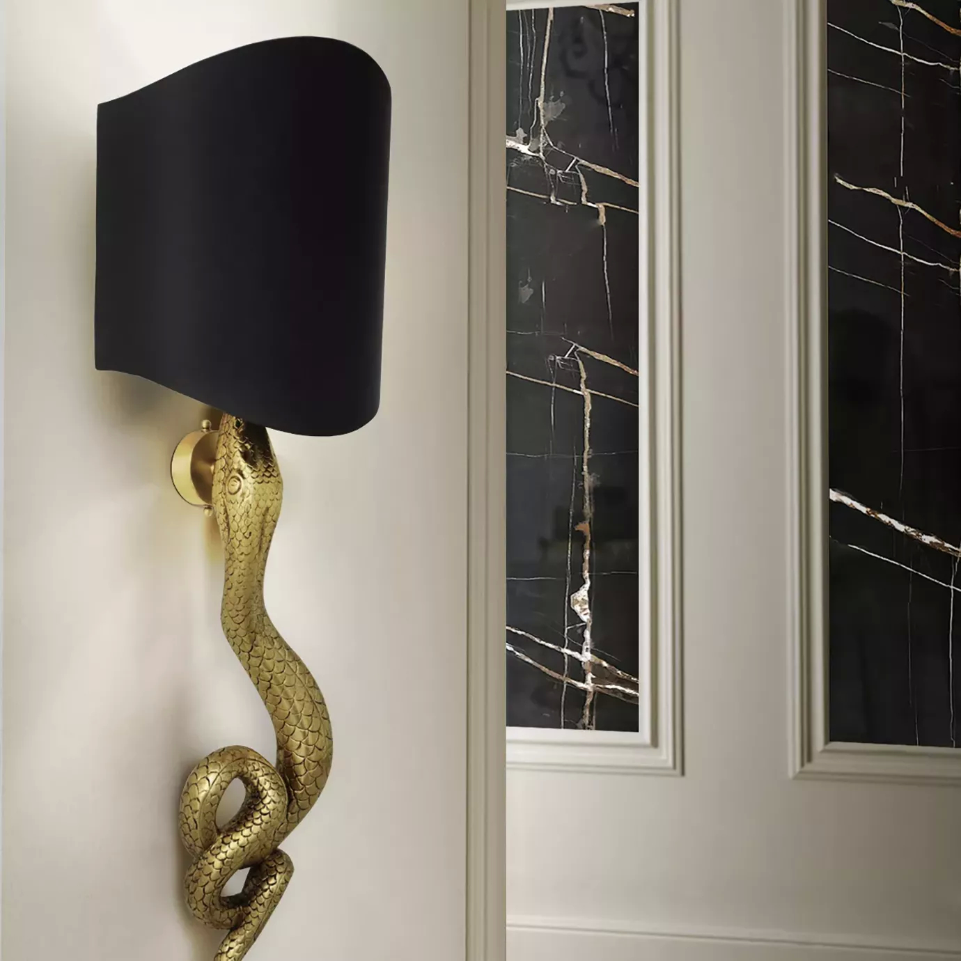 Serpentine Sconce with a gold snake base and black shade, mounted on a white wall next to dark marble panels.