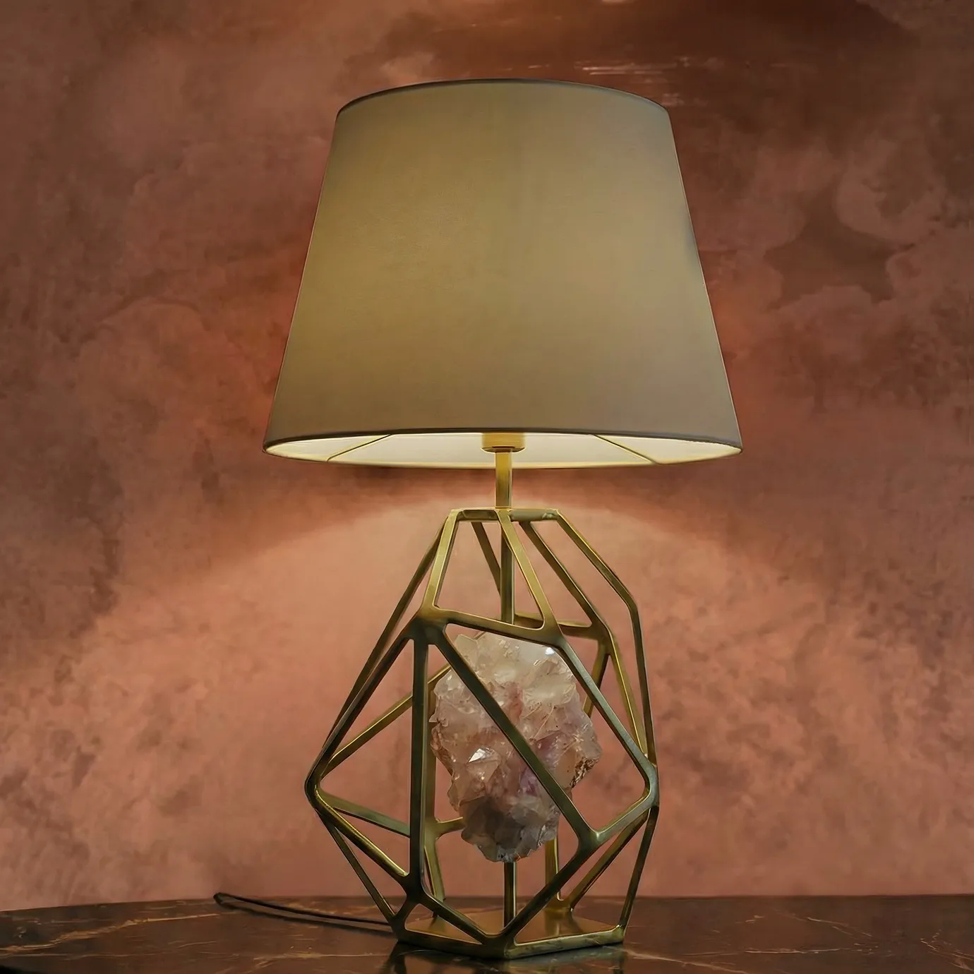 KOKET Gem Table Lamp with a geometric brass structure and illuminated natural crystal center, displayed on a marble console against a textured blush wall for a high-end interior décor setting.