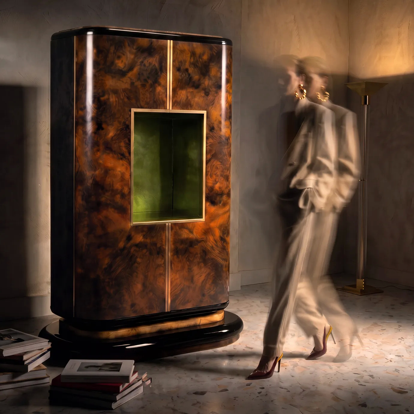 Eleanor armoire by KOKET in a dark interior with a person walking past, books on the floor and the green niche lit.