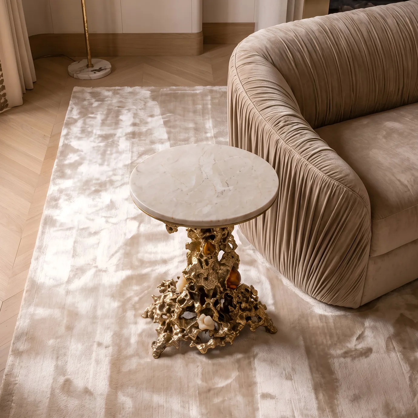 Empress side table by KOKET beside a pleated velvet sofa on a silver rug, cream marble top on a sand-cast brass base.