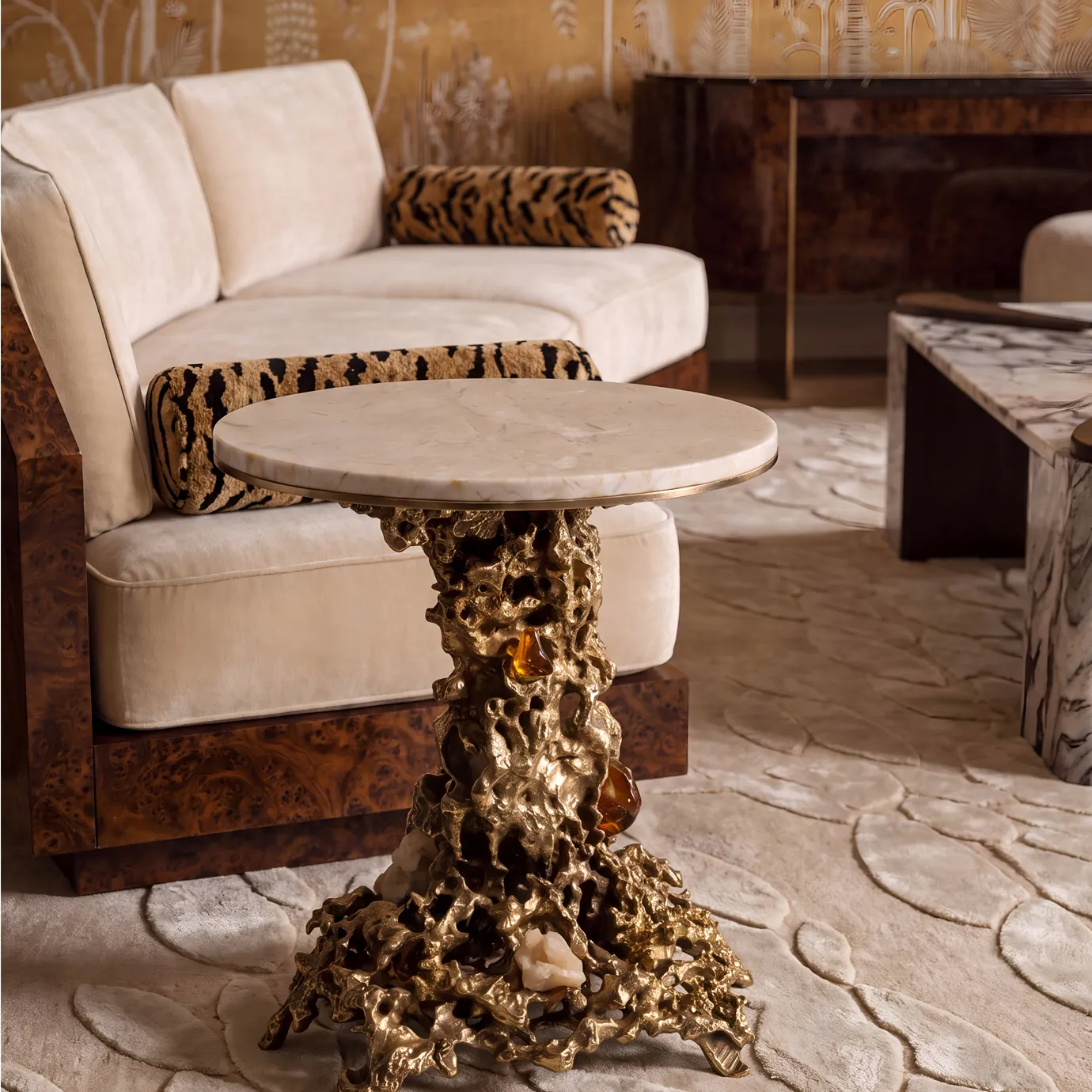 Empress side table by KOKET by a cream velvet sofa and palm-motif wallcovering, cream marble top on a sand-cast brass base.