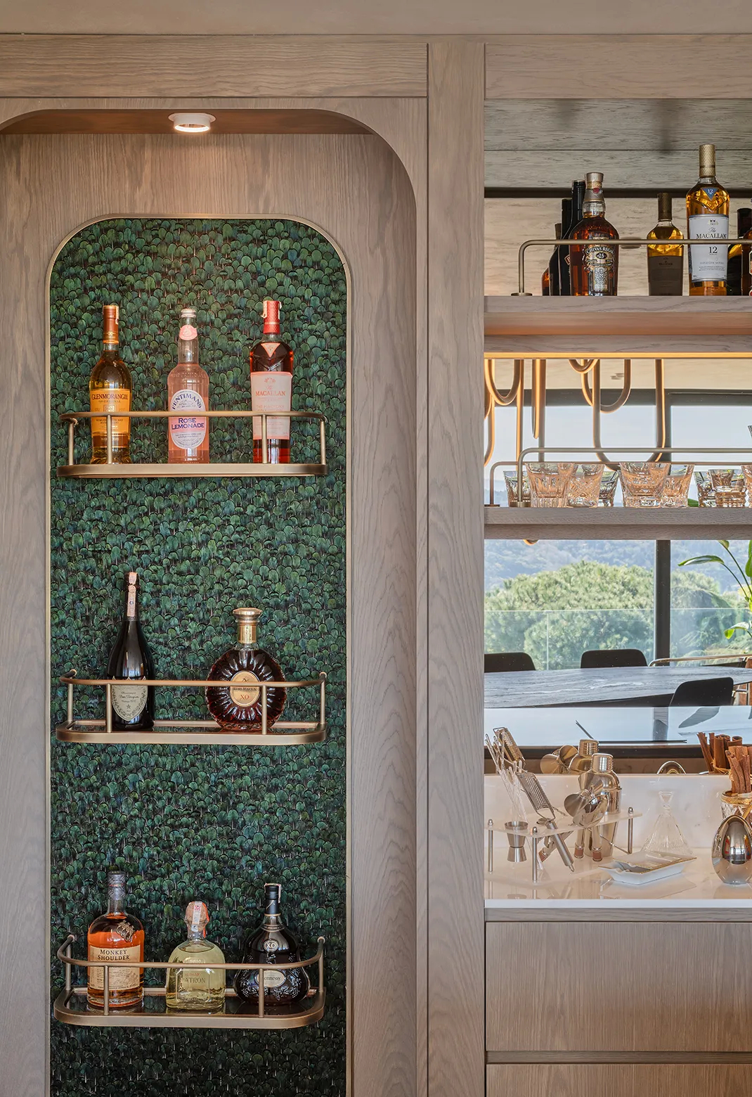 Detail of a luxury bar with brass shelves and KOKET’s iridescent peacock feather wallpaper as a bold, opulent backdrop.