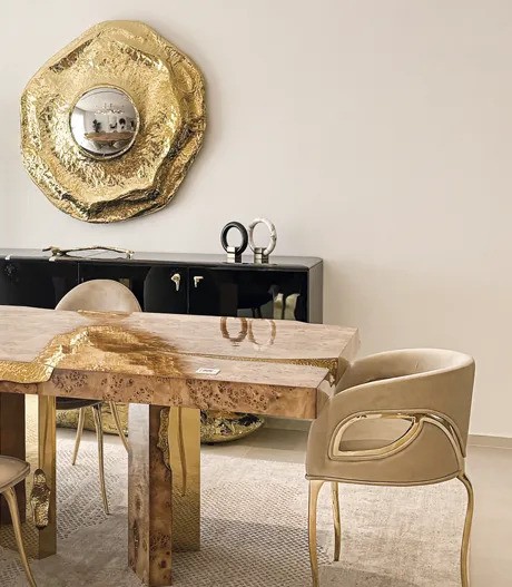 A luxurious dining room featuring a sleek, black sideboard and an ornate gold mirror. A dining table with a glossy, light wood top is surrounded by plush, beige Chandra Dining Chairs with gold accents.