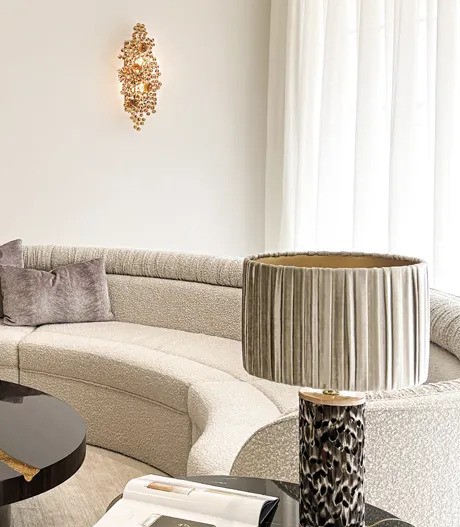 A living room featuring luxury furniture. A curved sofa sits below an Eternity Amber Glass Sconce and next to a Tailor Folie Feather Table Lamp.